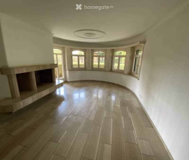 Empty room with wooden flooring, white walls, large windows, and a fireplace on the left side.