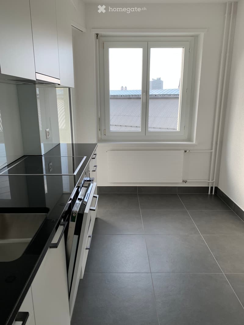 Empty kitchen with dark grey tiled floor, white walls, and a large window overlooking a rooftop. Minimalist design with a black countertop and white cabinets visible on the left side.