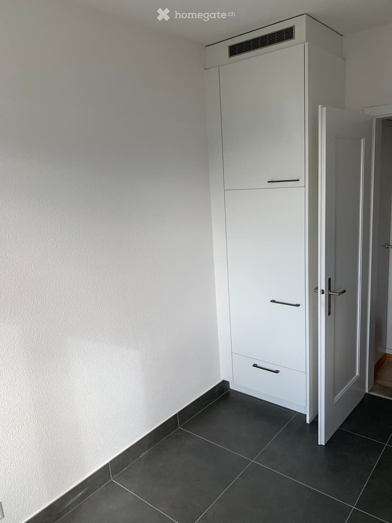 A narrow hallway with a white cabinet and a door leading to another room. The floor is tiled in dark gray, and the walls are painted white.