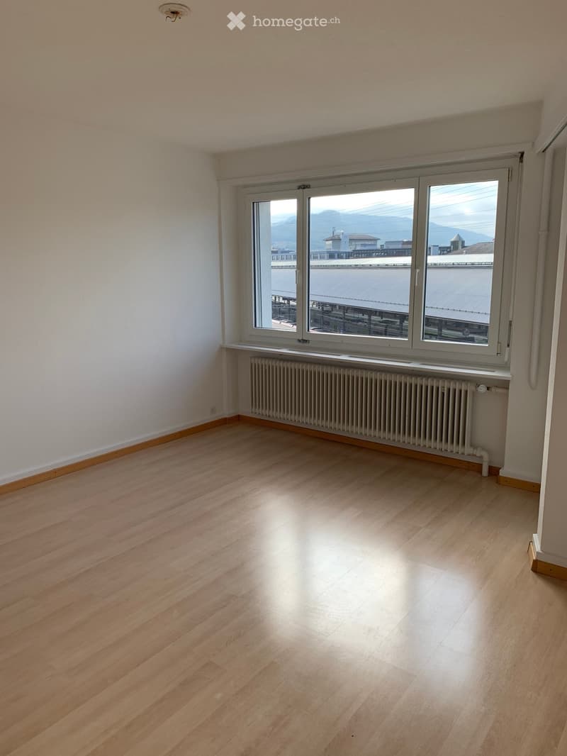 Empty room with wooden flooring, white walls, and a large window offering a view of the city and mountains. A radiator is placed under the window.