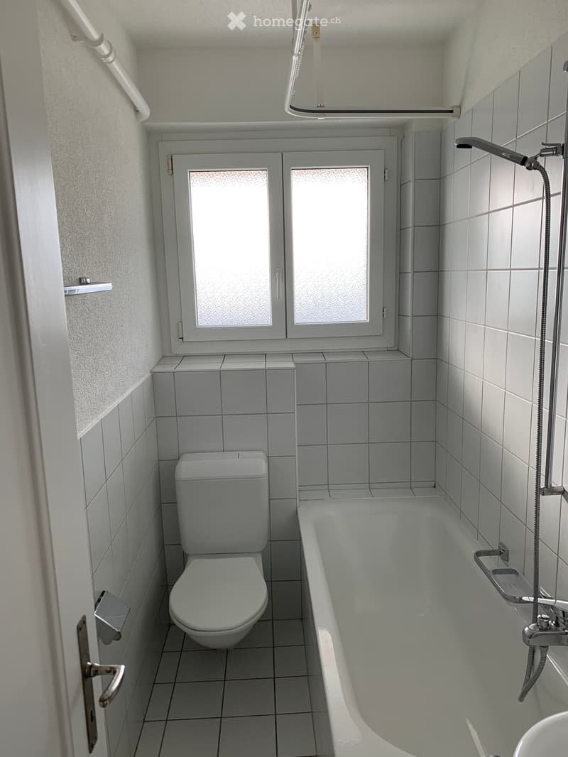 Modern bathroom with white tiled walls and floor, featuring a toilet, bathtub, and a window with frosted glass panes.