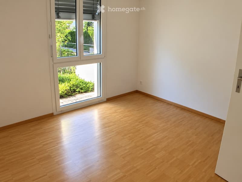 Empty room with light wooden flooring and white walls, featuring a large window and sliding glass door leading to a garden area.
