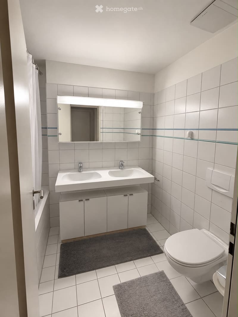 Modern bathroom with white tiles, double sink, large mirror, toilet, and two grey rugs.