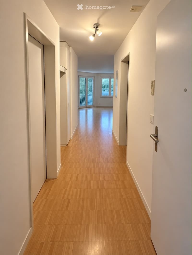 A clean, well-lit hallway with wooden flooring, white walls, and recessed ceiling lighting. The hallway features several doors and leads to a room with large windows and sliding glass doors.