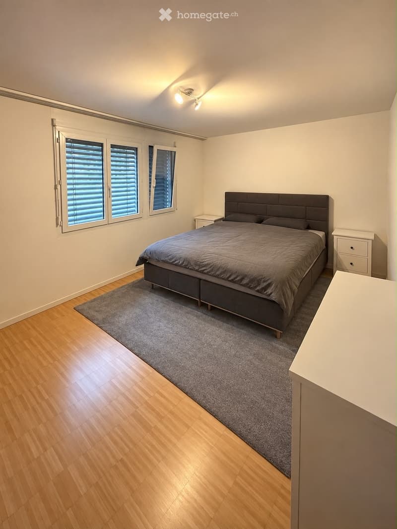 Modern bedroom with wooden flooring, large windows with blinds, a bed with dark headboard and grey bedding, a nightstand, and a grey rug.