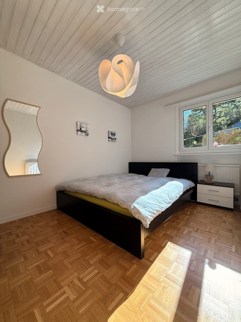 Cozy bedroom with wooden herringbone flooring, large bed with dark headboard, nightstand with lamp, wall-mounted mirror, and decorative picture frames.