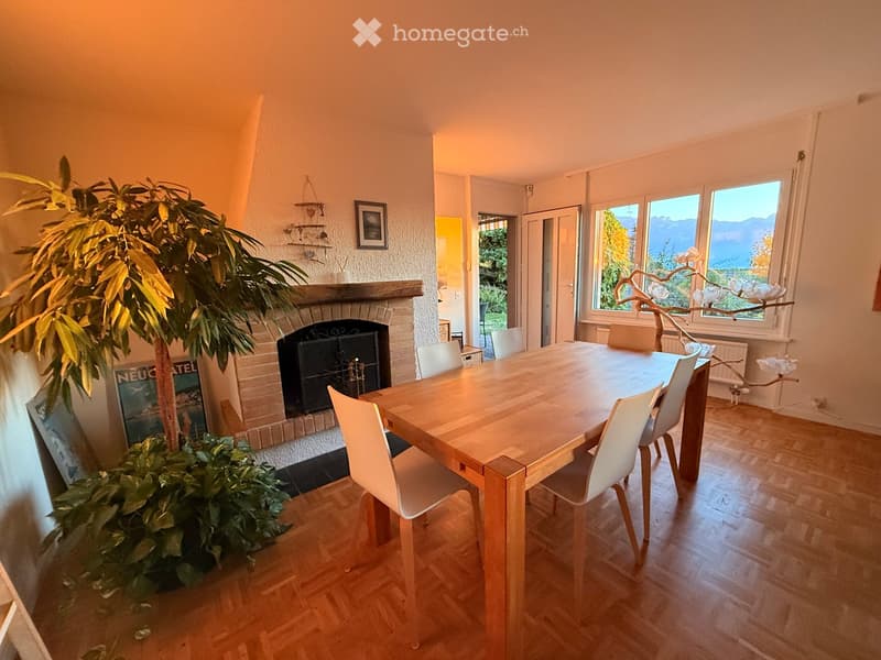 Dining room with a wooden dining table, white chairs, a brick fireplace, and large windows offering a scenic view.