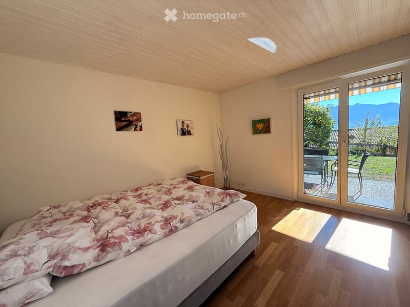 Bright bedroom with wooden flooring, large bed with floral duvet, nightstand, chair, and framed pictures on the wall. Large sliding glass doors open to a patio with outdoor furniture and a scenic mountain view.