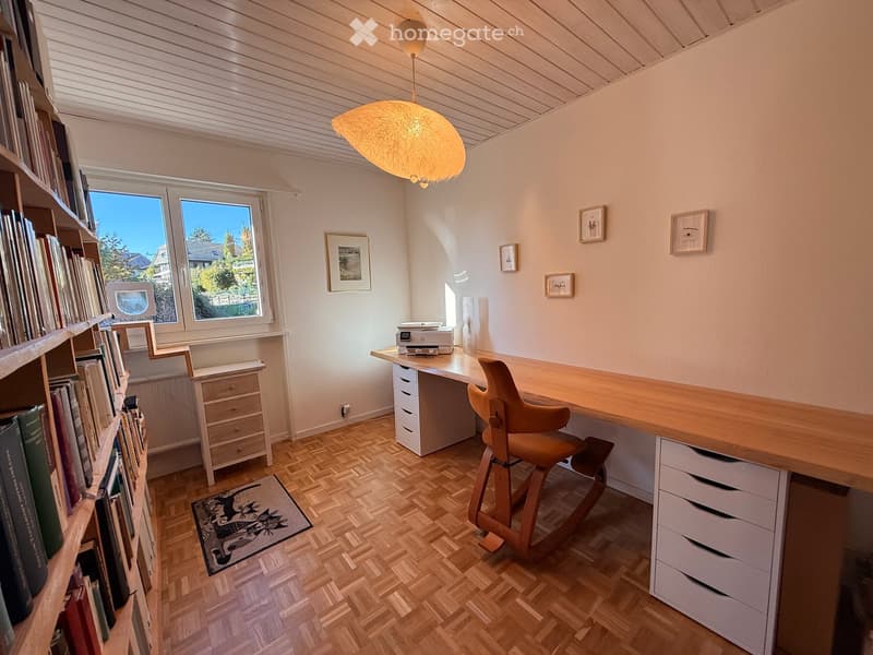 A well-lit study or home office with wooden flooring, a large wooden desk, a comfortable chair, a bookshelf filled with books, a printer, and several picture frames on the walls.