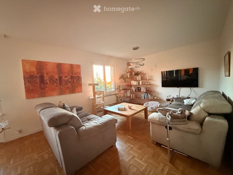 Bright living room with wooden flooring, a grey sectional sofa, a coffee table, a cat tree, a bookshelf, and a television mounted on the wall.