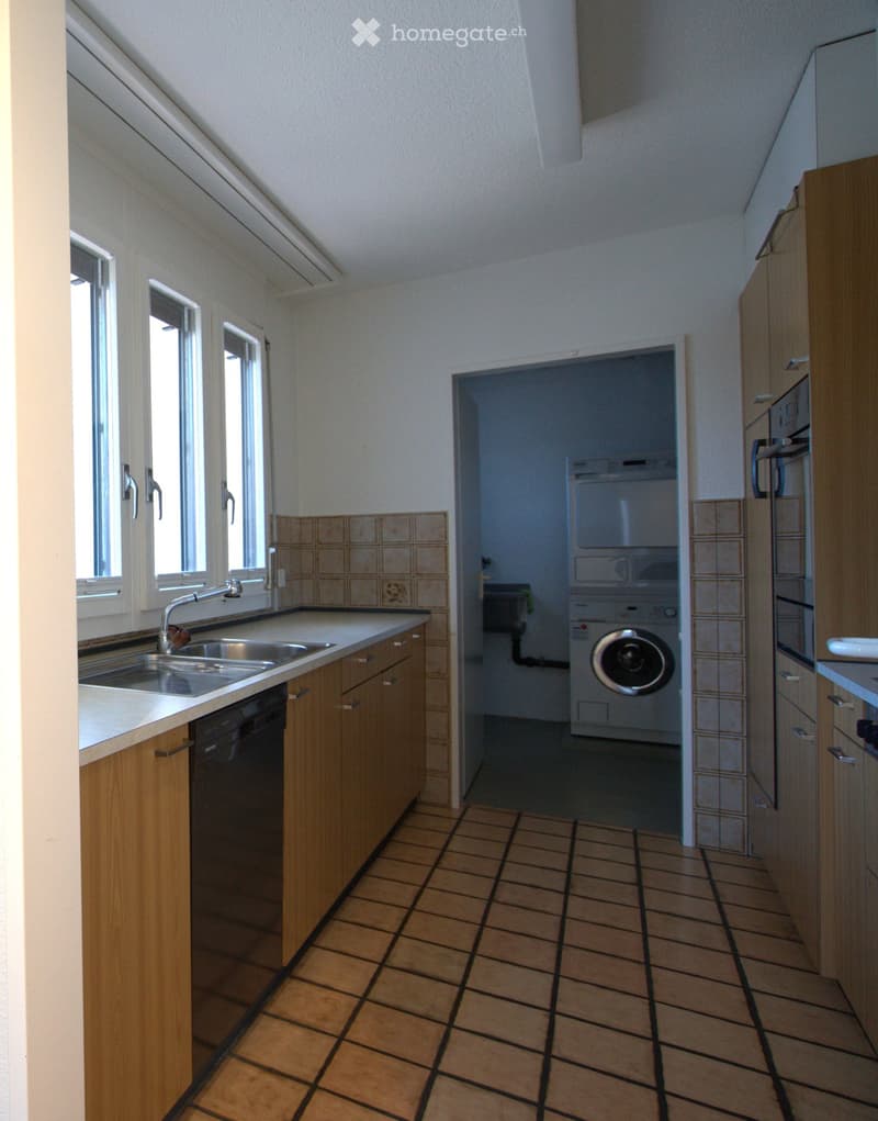 Bright kitchen with wooden cabinets, tiled floor, stainless steel sink, and dishwasher. Laundry area visible with washing machine.