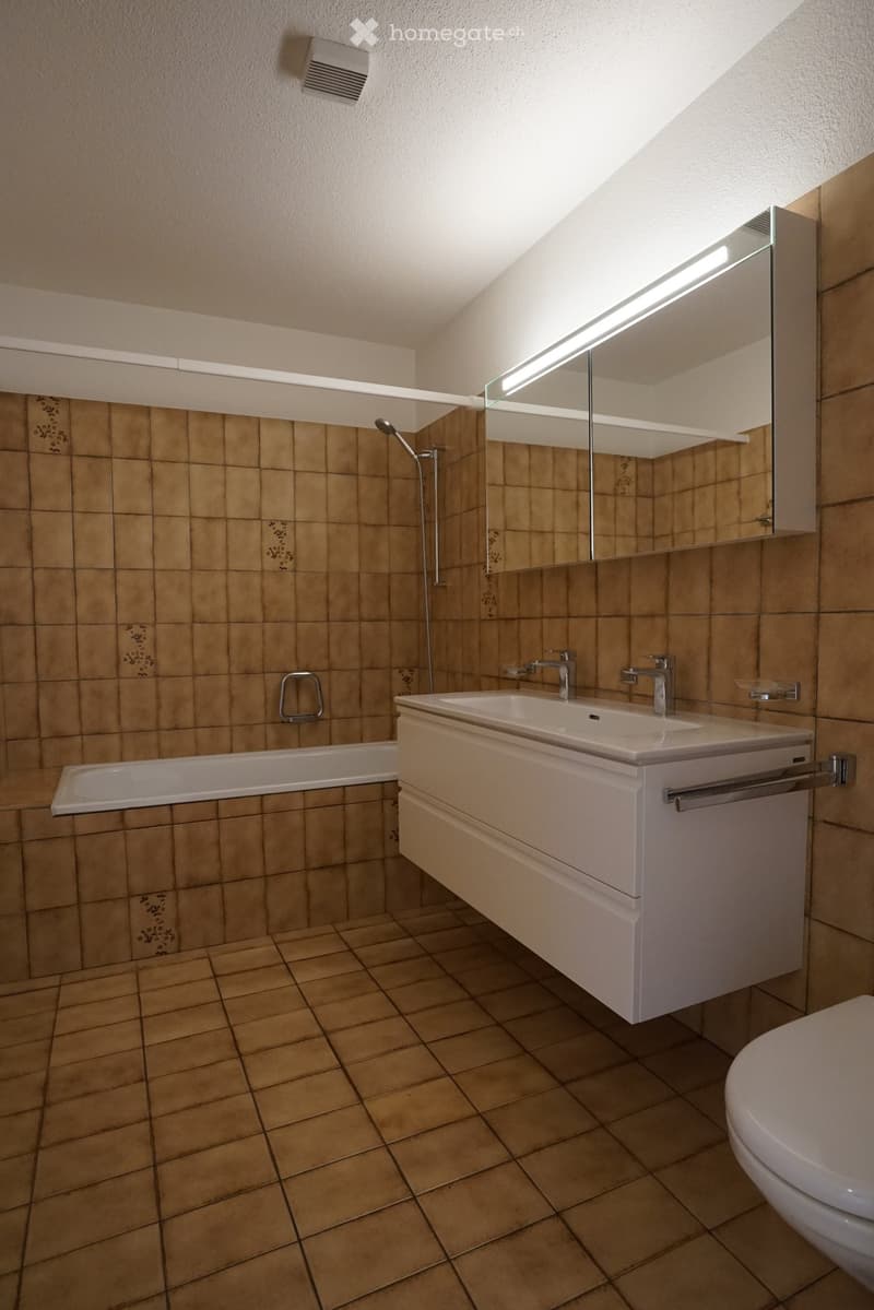 Modern bathroom with beige tiles, a bathtub, a double sink with modern faucets, a large mirror, and a toilet.