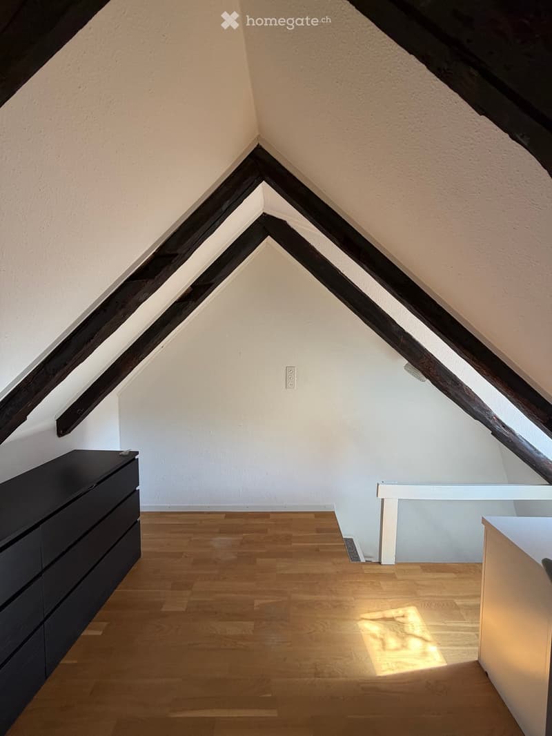 Attic room with wooden floor, slanted wooden beams, white walls, and a small black cabinet and white table.