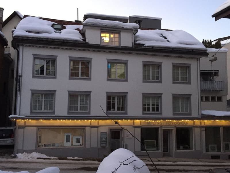 Three-story building with snow-covered roof and illuminated storefront sign reading "M. LINSI FOTOGRAF." Windows on upper floors are lit, and the ground floor has glass display cases.