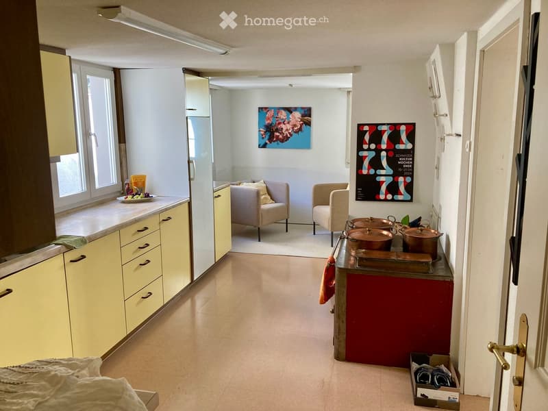 Open-plan kitchen and living area with light yellow cabinets, a red kitchen island, two beige armchairs, a refrigerator, and various decorative items including a painting and a poster.