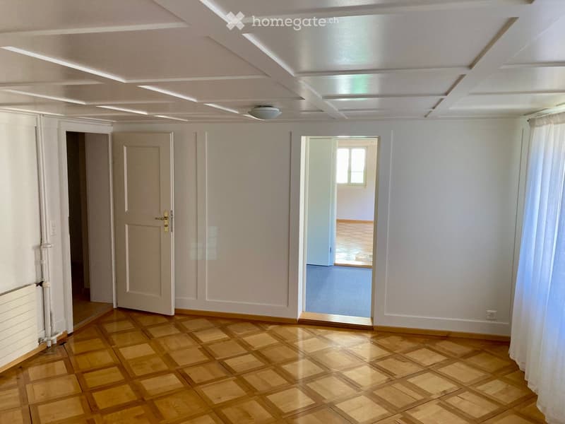 Empty room with wooden parquet flooring, white walls, and a white ceiling with recessed lighting. Features include a radiator, a door, and a window with a curtain.