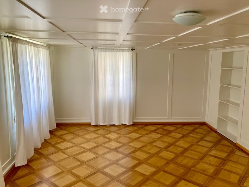 Empty room with wooden parquet flooring, white walls, and built-in shelving unit. White curtains cover the windows, and the ceiling has recessed lighting.