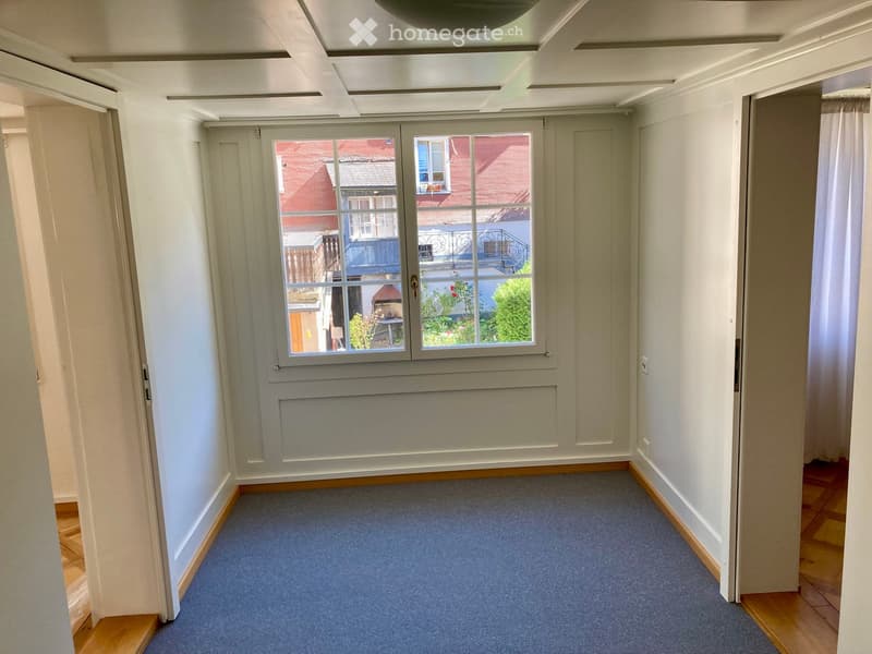Empty room with light-colored walls, carpeted floor, and a large window showing a view of neighboring buildings and greenery.