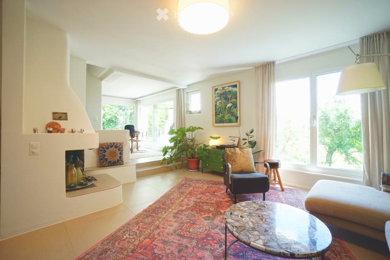 Bright living room with large windows, fireplace, sofa, coffee table, armchair, and decorative plants.