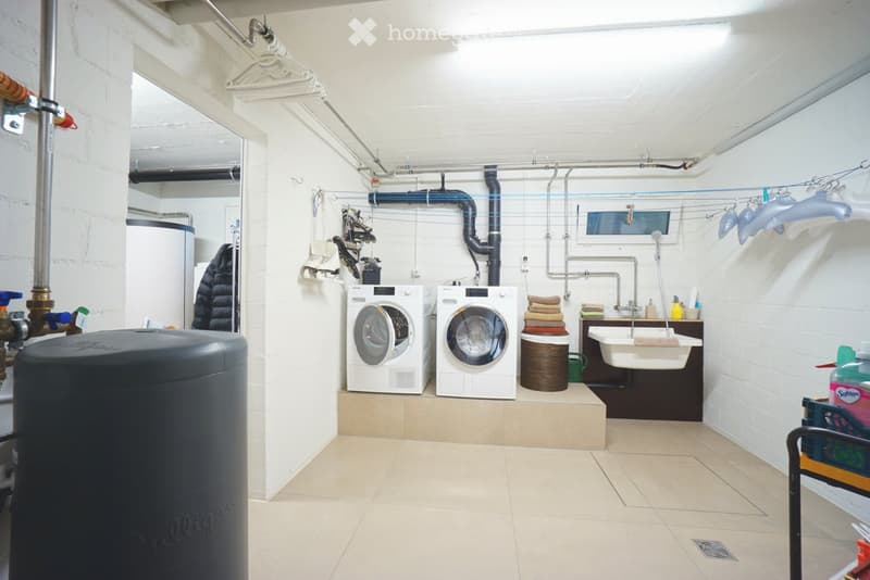 Laundry room with white tiled floor, white brick walls, two washing machines, a sink, and a laundry basket.
