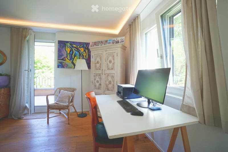 Brightly lit room with wooden flooring, a white desk with a computer setup, a wicker chair, a wooden cabinet, and a large window with curtains.