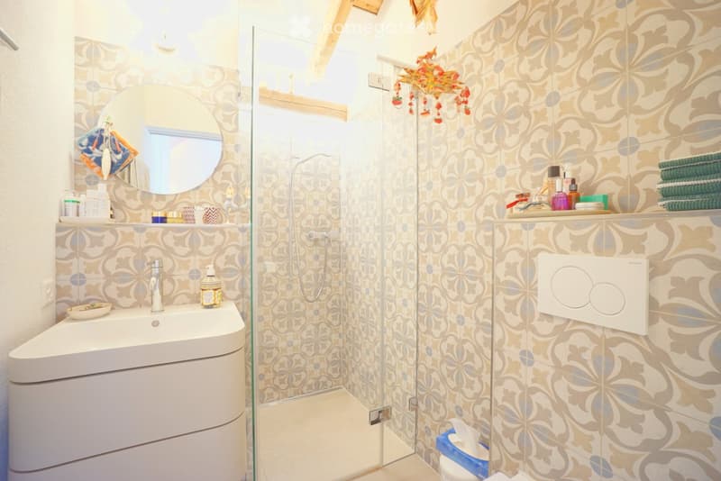 Modern bathroom with a white sink, round mirror, glass shower enclosure, and decorative floral tiles. Includes towels, toiletries, and a light fixture.