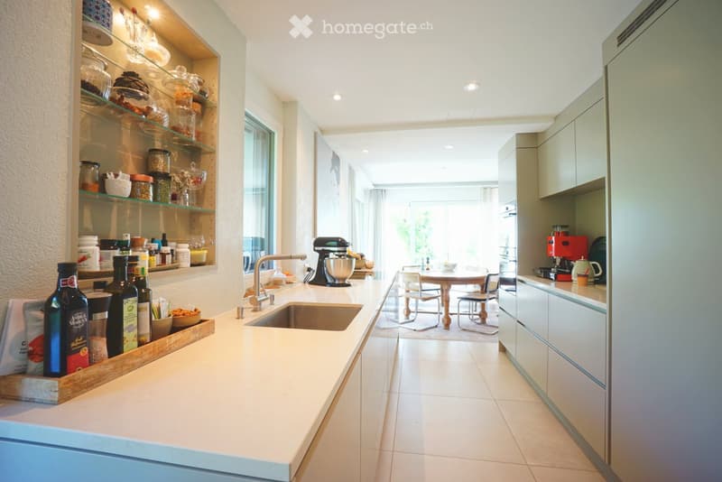 Modern kitchen with white countertops, stainless steel sink, mixer, coffee maker, and open shelving displaying jars and bottles. A small dining area with a table and chairs is visible in the background.