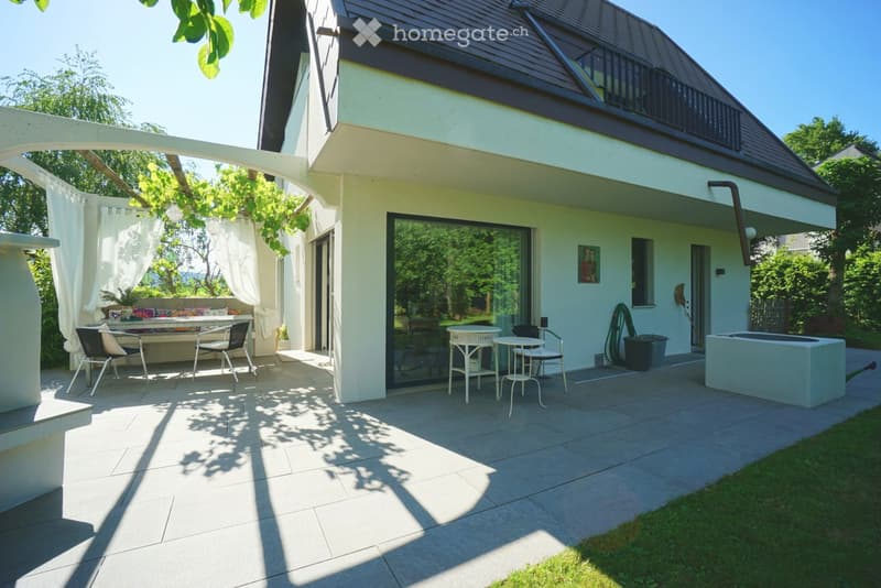 A modern patio area with a tiled floor, a small seating area with a table and chairs, a sofa with cushions, and a small round table with stools. The patio is attached to a house with a glass door leading inside.
