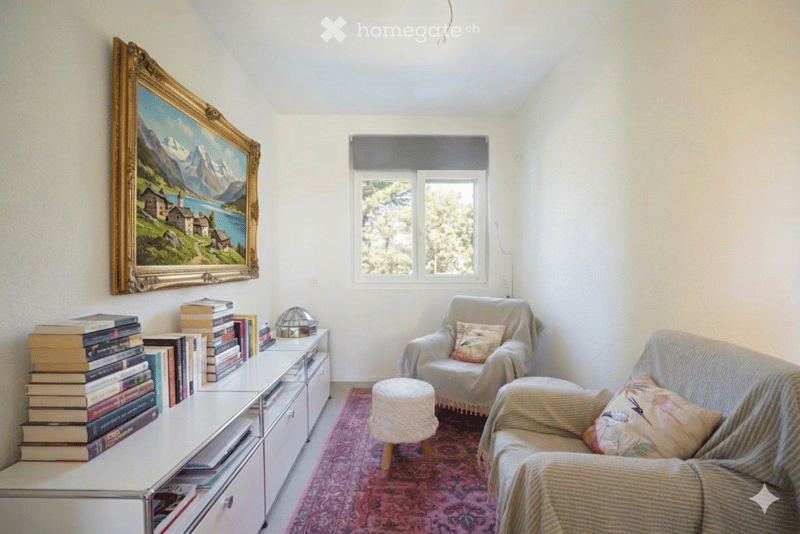 A cozy living room with a large window, a sofa, armchair, small round stool, a side table with books, and a framed painting on the wall.