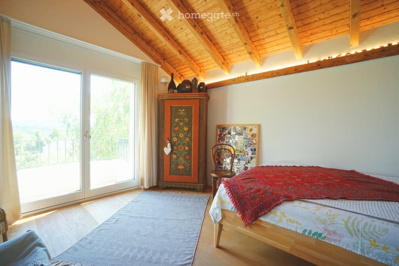 Cozy attic bedroom with wooden slanted ceilings, large window offering a scenic view, a bed with a red throw, a wooden chair, a colorful cabinet, and a small table with decorative items.