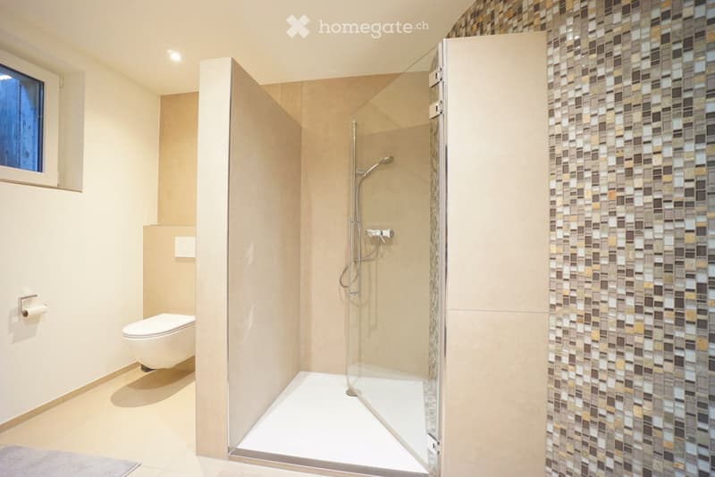 Modern bathroom with a toilet, glass-enclosed shower, and mosaic tile accent wall. Features a sleek, minimalist design with warm beige tones and a small window providing natural light.