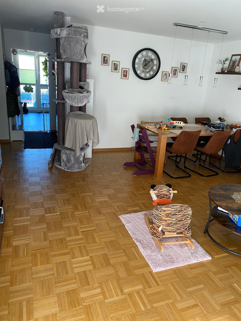 A well-lit room with wooden flooring, a cat tree, a dining table with chairs, a toy on a rug, and a large wall clock.