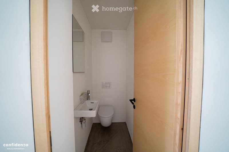 Compact bathroom with white walls, a sink, toilet, and a mirror. The space features light wooden door frames and a dark floor.