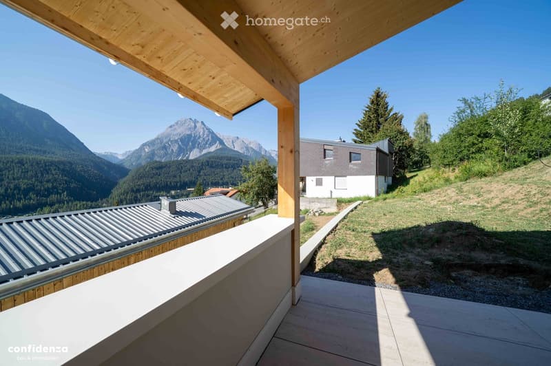 Balcony with wooden beams and a metal roof, overlooking a scenic mountain landscape with a modern house in the background.