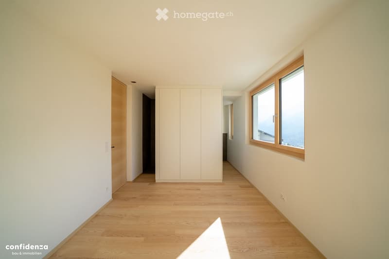 Empty room with light wooden flooring, white walls, and large windows offering a scenic view. The space includes built-in white cabinetry and a wooden door leading to another area.