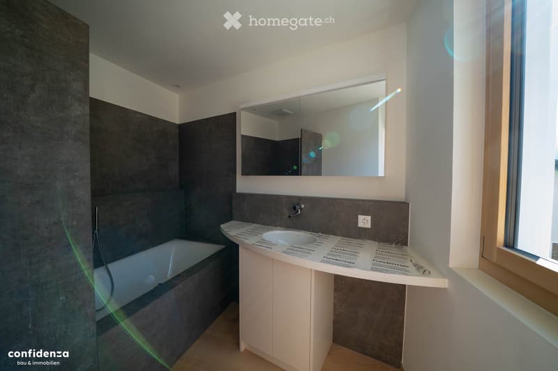 Modern bathroom with a bathtub, sink, and large mirror above the sink. The walls are tiled in dark gray, and the floor is wooden.