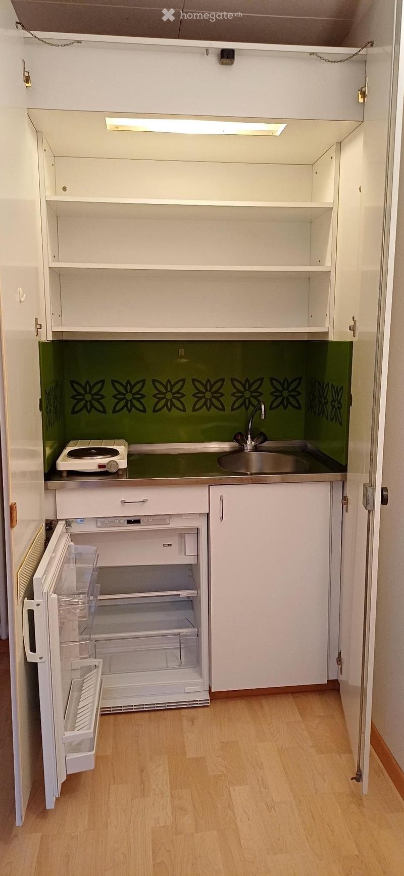 Compact kitchen with white cabinets, a small refrigerator, a sink with a faucet, and a stove, featuring a green backsplash with floral designs.