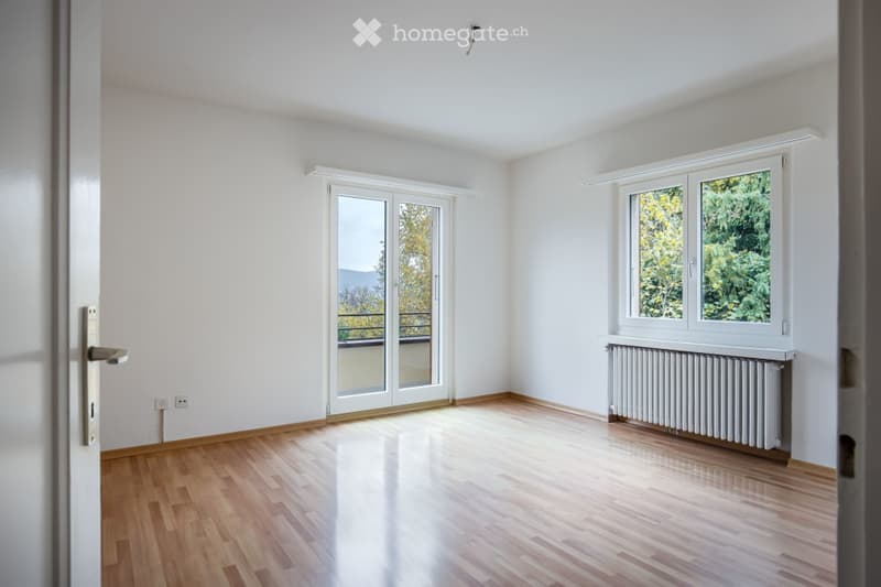 Empty room with wooden flooring, white walls, large windows, and a sliding glass door leading to a balcony.