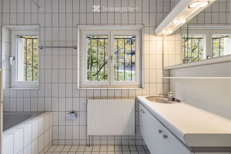 Bright bathroom with white tiled walls and floor, featuring a sink with a modern faucet, a bathtub, a radiator, and large windows with white frames. A mirror with built-in shelves is mounted on the wall above the sink.