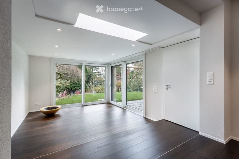 Bright, minimalist room with wooden flooring, large glass sliding doors opening to a garden, and a skylight above. A single decorative bowl sits on the floor.