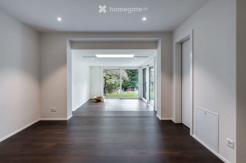 Modern, minimalist hallway with dark wooden flooring, white walls, recessed lighting, and a large opening leading to a garden area.