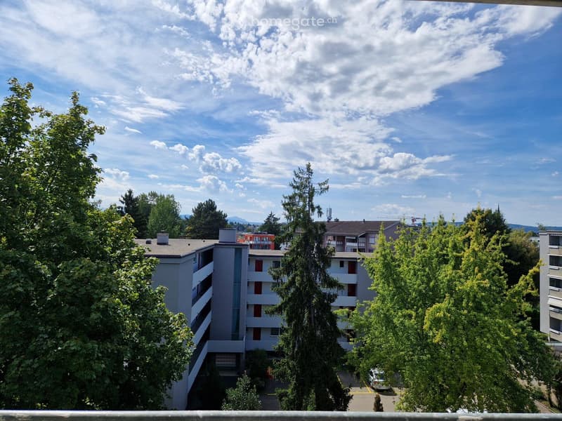 Balcony view of a multi-story residential building surrounded by trees, with a clear blue sky and scattered clouds in the background.