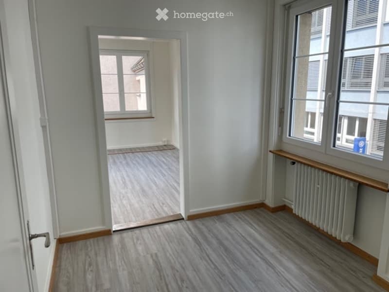 Empty room with light grey walls, wooden flooring, and a window with a radiator below. An open doorway leads to another room with similar flooring and a window.