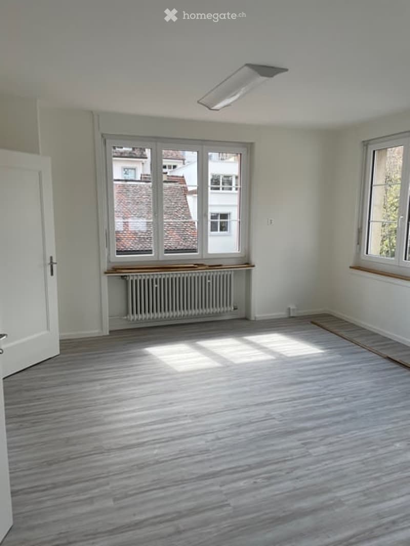 Empty room with light-colored wooden flooring, white walls, large windows with wooden sills, and a radiator below one window. The room has a simple, clean design with no furniture.