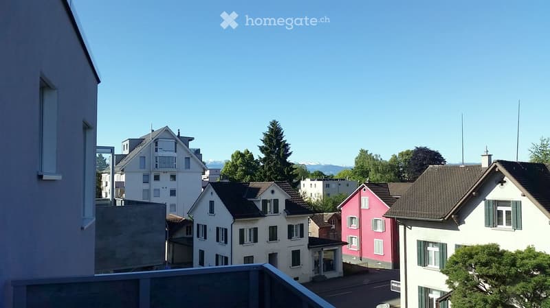 Balcony view overlooking a residential neighborhood with multi-story houses, trees, and distant mountains under a clear blue sky.