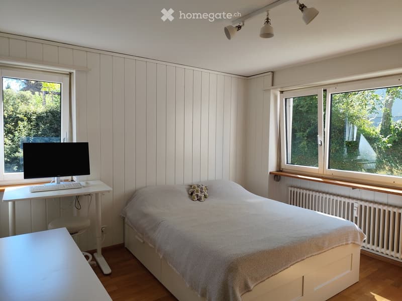 Cozy bedroom with a single bed, a desk with a computer, and a chair. The room features wooden flooring, white paneled walls, and large windows with a view of greenery.
