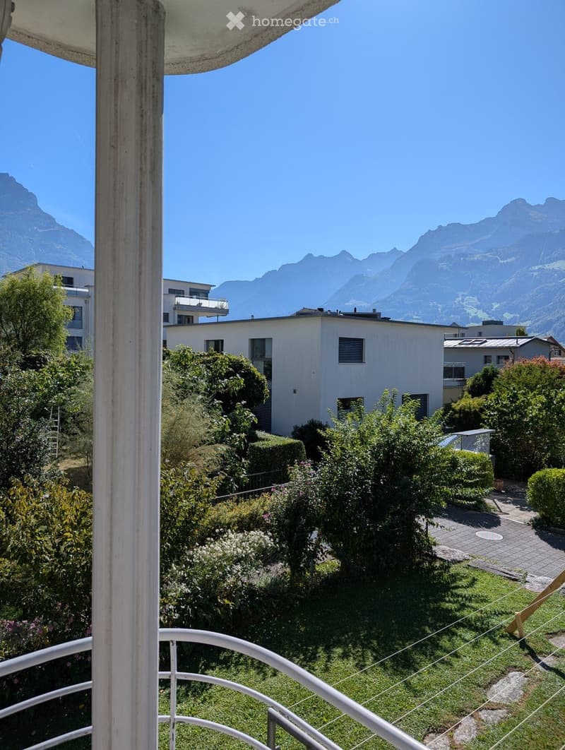 Balcony overlooking a garden area with greenery, a paved pathway, and distant mountain views.