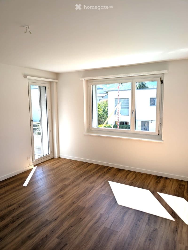 Empty room with wooden flooring, white walls, large windows, and a glass door leading outside.