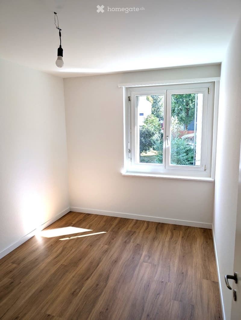 Empty room with light-colored walls, wooden flooring, and a single window with white frame. Natural light streams in, creating a bright and minimalist space.