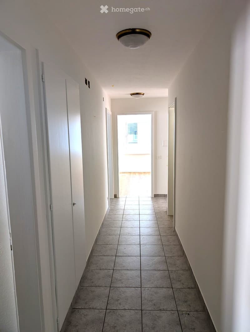 Empty hallway with tiled floor, white walls, and several doors leading to other rooms. Natural light streams in from an open doorway at the end.
